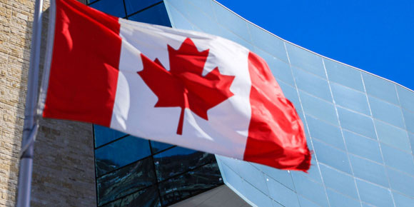 Drapeau canadien flottant au vent devant le Musée canadien pour les droits de la personne; on aperçoit un coin de ciel bleu vif.