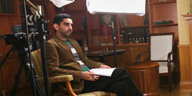 Man sitting with a pen and paper in a wood-paneled office, surrounded by lights and a video camera.