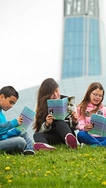 Quatre enfants lisant chacun un livre sur une colline gazonnée devant le Musée canadien pour les droits de la personne.