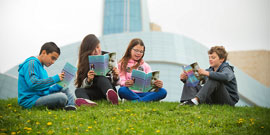 Four children each reading a book on a grassy hill in front of the Canadian Museum for Human Rights.